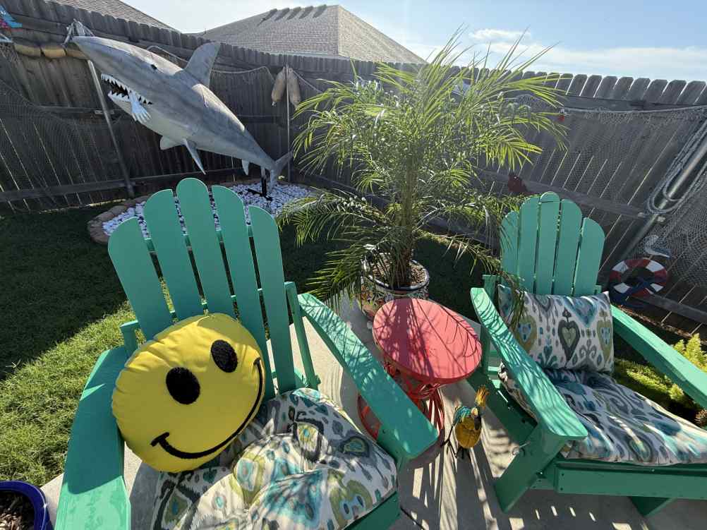 Close-up of the nine-foot rusty metal shark sculpture beside teal Adirondack chairs and a palm plant